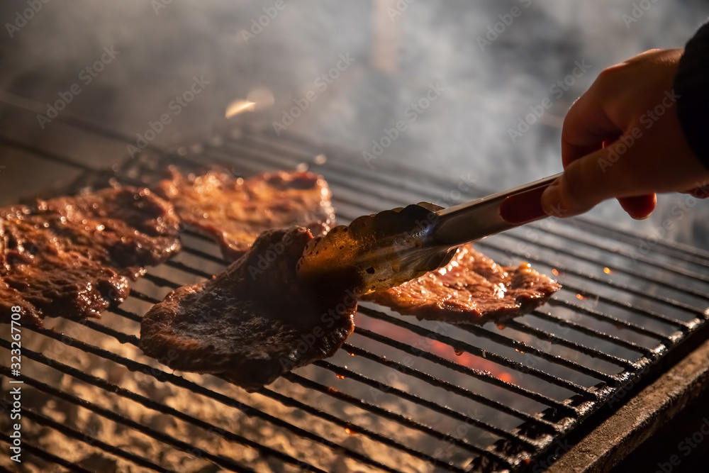 in the evening a barbecue grill on which tasty juicy steaks are grilled over an open fire, a hand holds tongs and turns the meat over. close-up, soft focus. smoke is highlighted in the evening a barbecue grill on which tasty juicy steaks are grilled over an open fire, a hand holds tongs and turns the meat over. close-up, soft focus. smoke is highlighted
