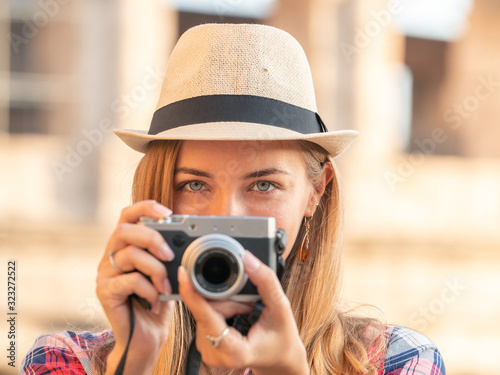 Young blonde woman with blue eyes and long hair, taking pictures with a vintage camera at Colosseum, Rome. Large hat, red squared shirt. © WineDonuts