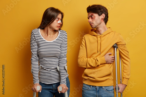 Dissatisfied woman and man have accident, unlucky day, have talk with eah other, stand on crutches, discuss recovery period, have wounds and scratches on body, isolated on yellow background. photo