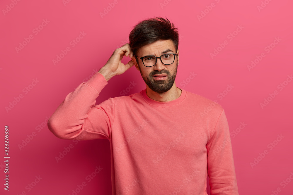 Serious bearded young man scratches head, thinks deeply about how to solve problem, makes decision, wears optical glasses, poses indoor against rosy background, makes up plan, has bad memory Serious bearded young man scratches head, thinks deeply about how to solve problem, makes decision, wears optical glasses, poses indoor against rosy background, makes up plan, has bad memory
