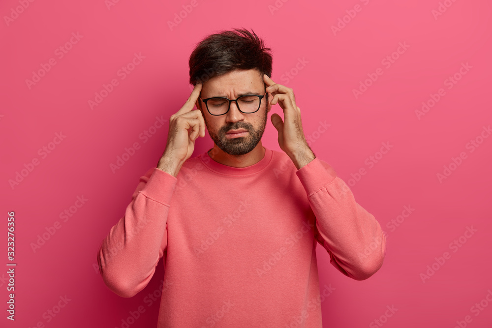 Stressed man rubs temples, has headache, closes eyes to relieve pain, suffers from migraine after noisy atmosphere, has thick beard, wears rosy jumper, stands against vibrant crimson background Stressed man rubs temples, has headache, closes eyes to relieve pain, suffers from migraine after noisy atmosphere, has thick beard, wears rosy jumper, stands against vibrant crimson background