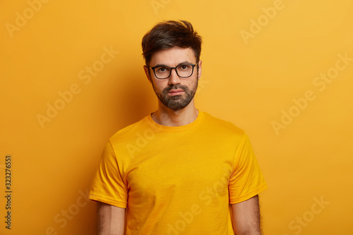 Half length shot of handsome serious man with beard, looks directly at camera, wears glasses and yellow t shirt, thinks about something, poses indoor. Monochrome shot. Human facial expressions