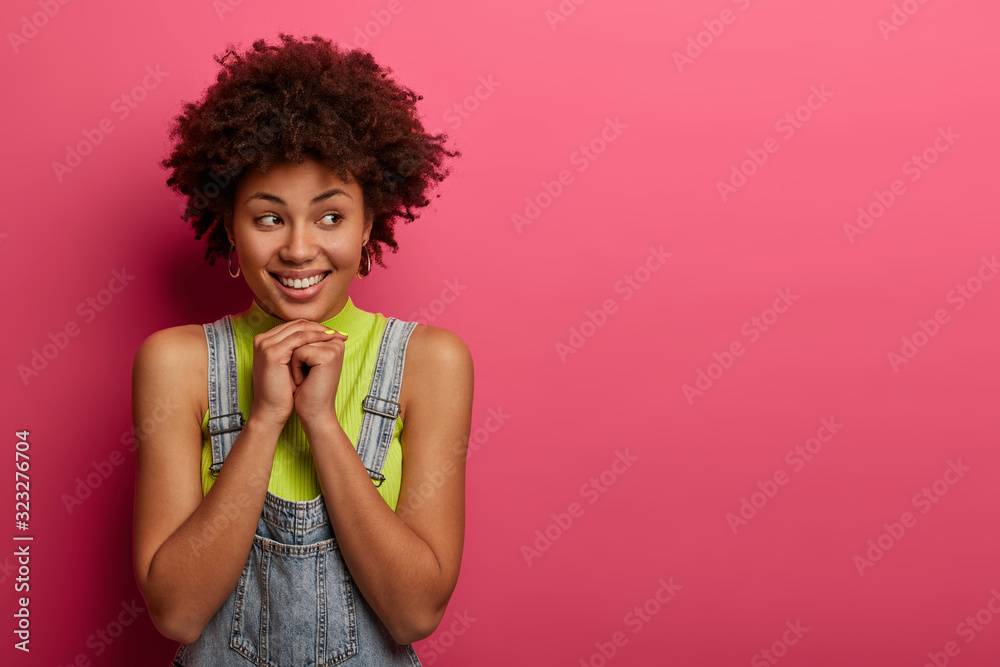 Lovely Afro American feminine model grins from joy, smiles toothily, keeps hands under chin, dressed casually, looks aside on copy space, isolated over pink background. People, emotions concept Lovely Afro American feminine model grins from joy, smiles toothily, keeps hands under chin, dressed casually, looks aside on copy space, isolated over pink background. People, emotions concept