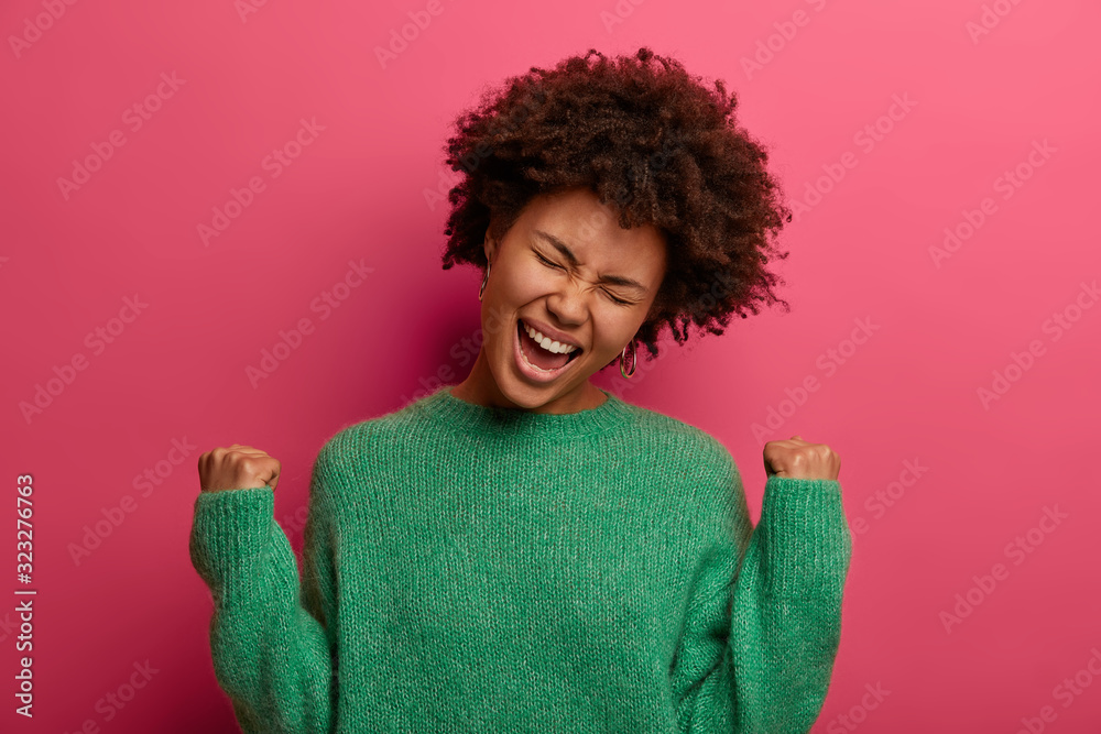 Ecstatic curly young woman exclaims and clenches fists with joy, feels like winner, triumphs and celebrates success, wears green sweater, poses against crimson background, celebrates victory Ecstatic curly young woman exclaims and clenches fists with joy, feels like winner, triumphs and celebrates success, wears green sweater, poses against crimson background, celebrates victory