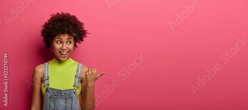 Happy girl asks you out, invites for drink, points thumb right, recommends promo or website, wears green t shirt and overalls, isolated over pink background, copy space for your advertising.