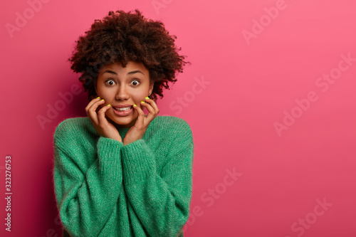 Photo of worried nervous woman clenches teeth  keeps hands on face  afraids of making mistake or speak in public  wears green sweater  models over rosy wall with copy space area. Negative emotions