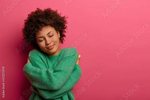 Cheerful young romantic woman expresses self love and care, tilts head and smiles gently, wears green oversized jumper, embraces own body, closes eyes, stands in studio against pink background photo