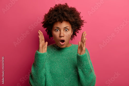 Photo of embarrassed curly stupefied woman shapes something very big, keeps mouth opened, explains size of big box, dressed in green sweater, isolated over pink background. Omg, its so huge!