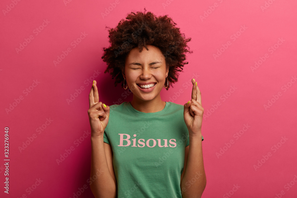 Happy curly African American woman with broad smile, shows white teeth, crosses fingers hopefully, dreams about getting proposal from boyfriend, wears t shirt, written word kisses, feels upbeat Happy curly African American woman with broad smile, shows white teeth, crosses fingers hopefully, dreams about getting proposal from boyfriend, wears t shirt, written word kisses, feels upbeat