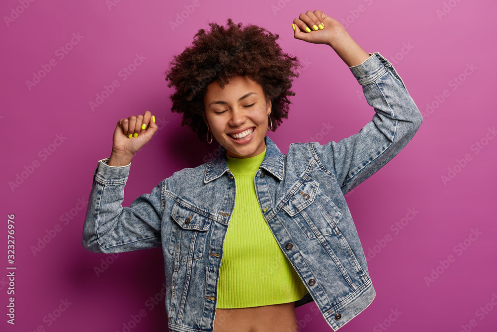 Carefree curly woman raises arms dances joyfully, has fun carried away with pleasant music, moves actively on disco floor, wears top and denim jacket, feels every tune of melody isolated on lilac wall Carefree curly woman raises arms dances joyfully, has fun carried away with pleasant music, moves actively on disco floor, wears top and denim jacket, feels every tune of melody isolated on lilac wall