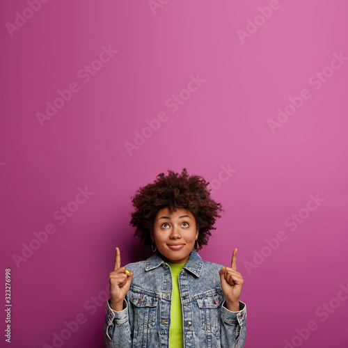 Curious lovely ethnic woman with curly hairstyle, points fore fingers upwards, wears denim jacket, promots object above, isolated over purple background, rejoices fall of prices, introduces copy space