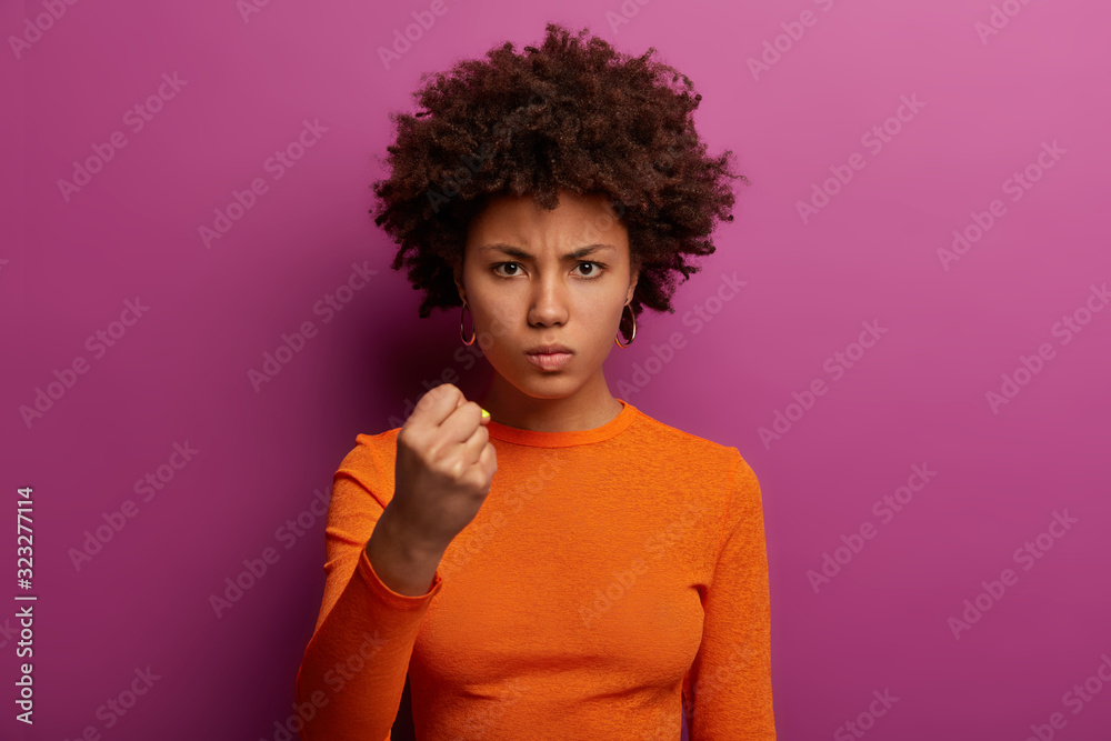 Aggressive outraged serious curly ethnic woman shows clenched fist, warns to revenge, going to punish coworkers for coming late at work, wears orange casual jumper, isolated on purple background