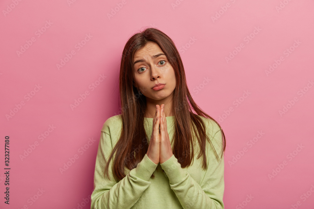 Portrait of imploring girl tilts head, purses lips, looks sadly at camera, makes praying gesture, asks for help and support, begs boyfriend for apologize, wears casual sweater, isolated on pink wall Portrait of imploring girl tilts head, purses lips, looks sadly at camera, makes praying gesture, asks for help and support, begs boyfriend for apologize, wears casual sweater, isolated on pink wall