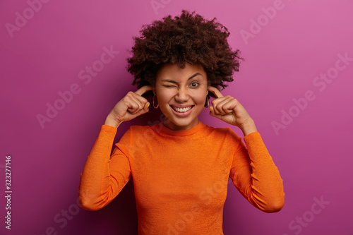 Cheerful curly haired woman feels relieved  plugs ear holes with index fingers  hears no sound or noise  shows white teeth  wears casual clothing  poses indoor. Body language  reaction  attitude