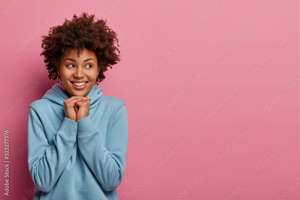Sincere positive dark skinned model holds hands under chin, being in good mood, wears blue sweatshirt, has happy smile on face, models against pastel rosy wall. People, emotions and lifestyle Sincere positive dark skinned model holds hands under chin, being in good mood, wears blue sweatshirt, has happy smile on face, models against pastel rosy wall. People, emotions and lifestyle