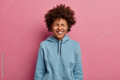 Εκτύπωση καμβά Portrait of overjoyed curly woman closes eyes and smiles broadly, shows white teeth, laughs while watches comedy, has upbeat mood, wears sweatshirt, isolated over pink background