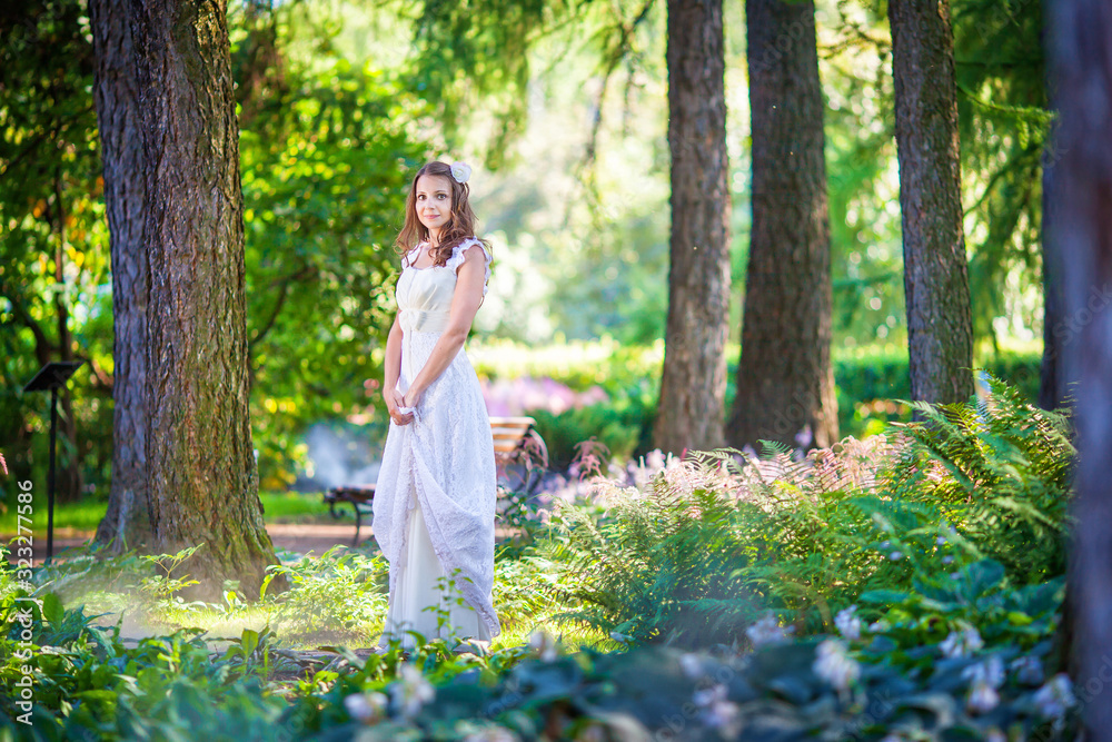 Pretty curvy brunette girl with long hair in white long dress standing in the green forest. Fairy princess stands on woods background. Portrait of young bride in park in sunny spring weather Pretty curvy brunette girl with long hair in white long dress standing in the green forest. Fairy princess stands on woods background. Portrait of young bride in park in sunny spring weather
