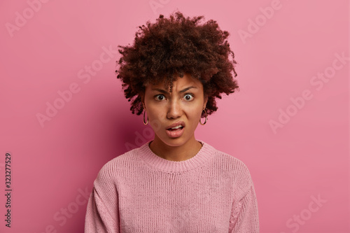 Portrait of angry displeased woman smirks face, expresses scorn and disdain, raises eyebrows, stares at camera, irritated by something, wears rosy sweater, expresses negative emotions and feelings