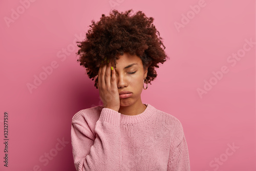 Portrait of dark skinned woman covers half of face, sighs from tiredness, has sleepless night working on project or diploma paper, feels sleepy during boring lecture at university, wakes up after nap photo