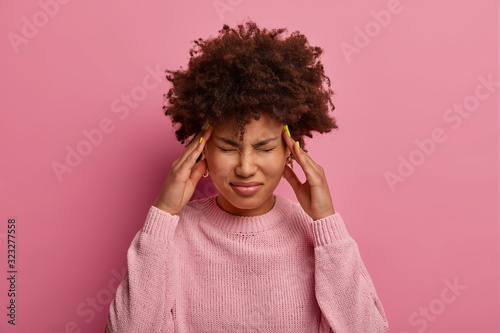Distressed dark skinned woman rubs temples, feels dizzy and severe headache, suffers from hangover after party, has problems with health, wears rosy sweater, grimaces from pain, compresses head photo