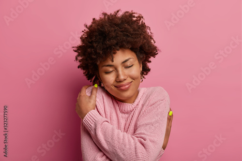 Self love and tenderness concept. Delighted gentle woman with curly hairstyle, embraces herself, hugs own body, closes eyes and wears warm soft casual jumper for cold weather, smiles cheerfully