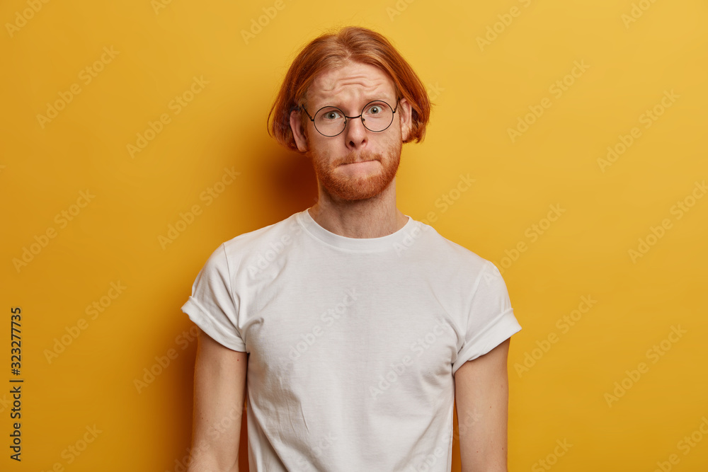 Handsome bearded man presses lips, looks with worried expressionn, tries to remember answer on question at exam, has trendy foxy hairstyle wears round glasses and casual outfit stands in yellow studio Handsome bearded man presses lips, looks with worried expressionn, tries to remember answer on question at exam, has trendy foxy hairstyle wears round glasses and casual outfit stands in yellow studio