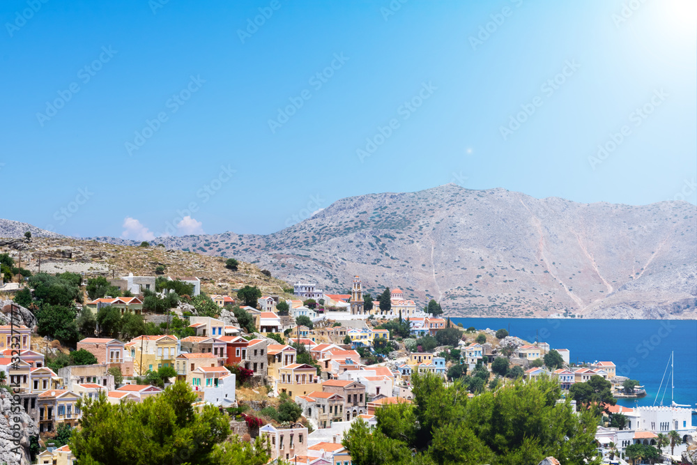 Picturesque view on tiny colorful houses on rocks, green trees near the Mediterranian sea on Greek island in sunny summer day with sunbeams, vacation on exotic islands