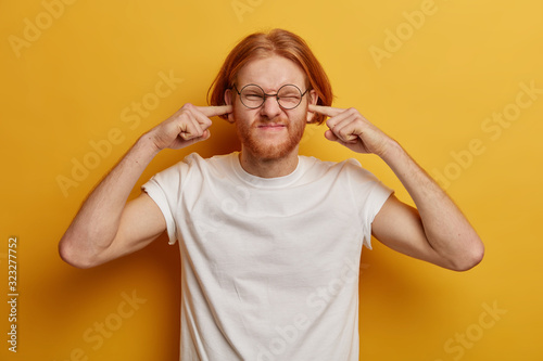 Disturbed ginger man plugs ears, ignores loud music, smirks face, hears unpleasant annoying sound, dissatisfied by noise, wears casual clothing, isolated on yellow wall. Noisy neighbours is problem