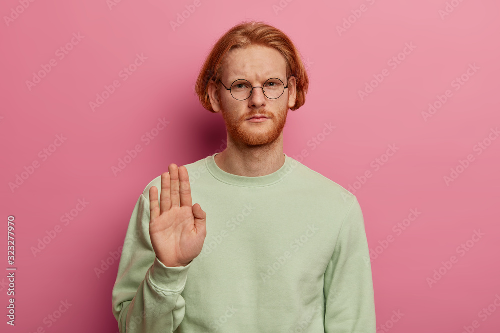 Serious bearded redhead man shows palm in stop gesture, expresses restriction and prohibition, looks with strict gaze at camera, dressed casually, isolated on pink background. Body language. Serious bearded redhead man shows palm in stop gesture, expresses restriction and prohibition, looks with strict gaze at camera, dressed casually, isolated on pink background. Body language.