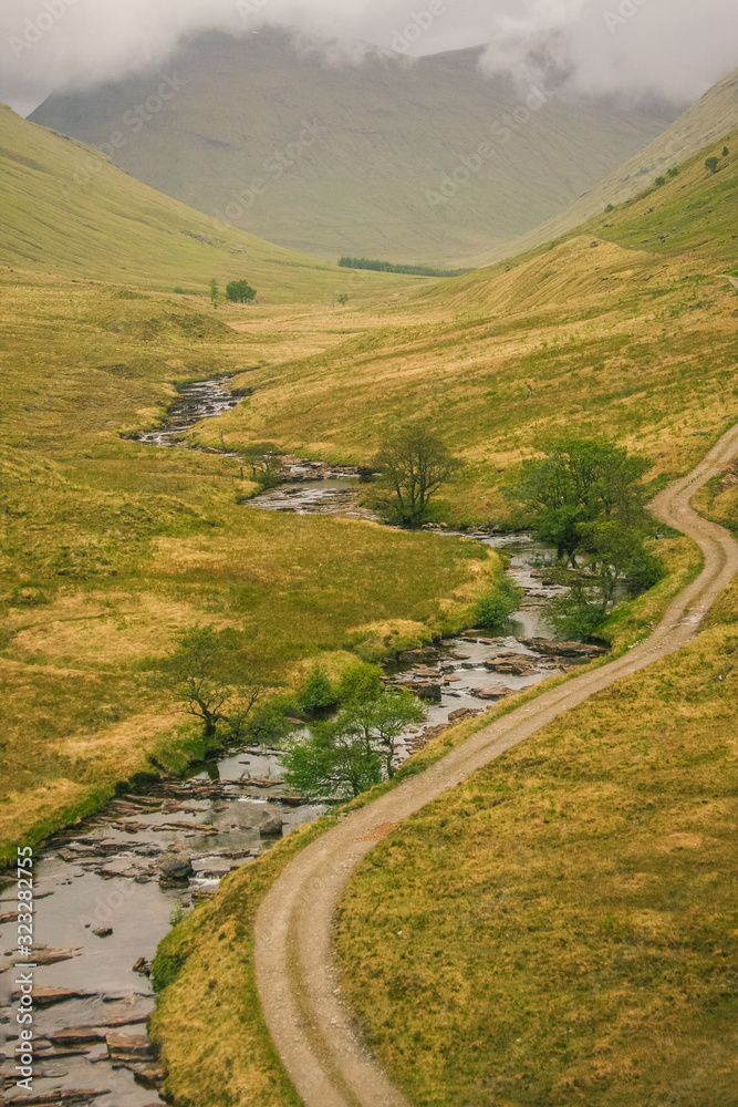 Vertical panorama of a typical scottish nature, with a fire road and a small river on a lush green meadow, with white clouds above.