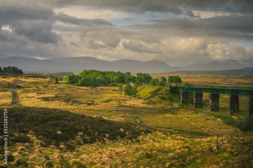 Obraz premium Green train bridge on a green scottish countryside in a foggy rainy weather. Hills and dense clouds in the distance, while traveling with a train.