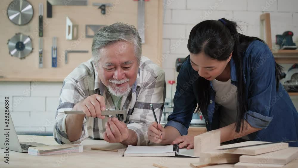 Asian senior male and teenage girl carpenter working design modern furniture in home workshop. Carpenter is high experience and presentation skill in lumber.