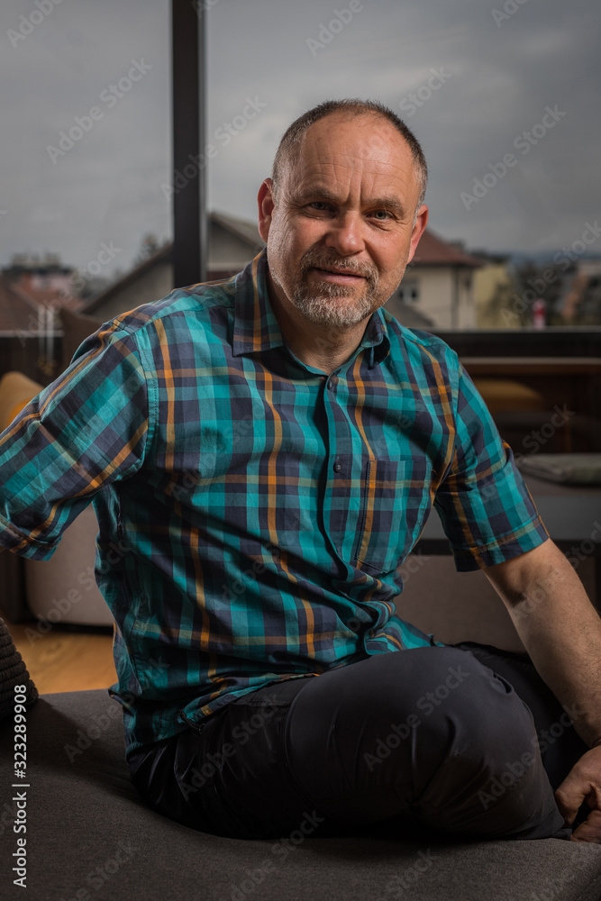 Fototapeta premium A caucasian man in his late forties with short hair and short gray beard is sitting in a couch and looking into the camera. Forest is seen behind the window.