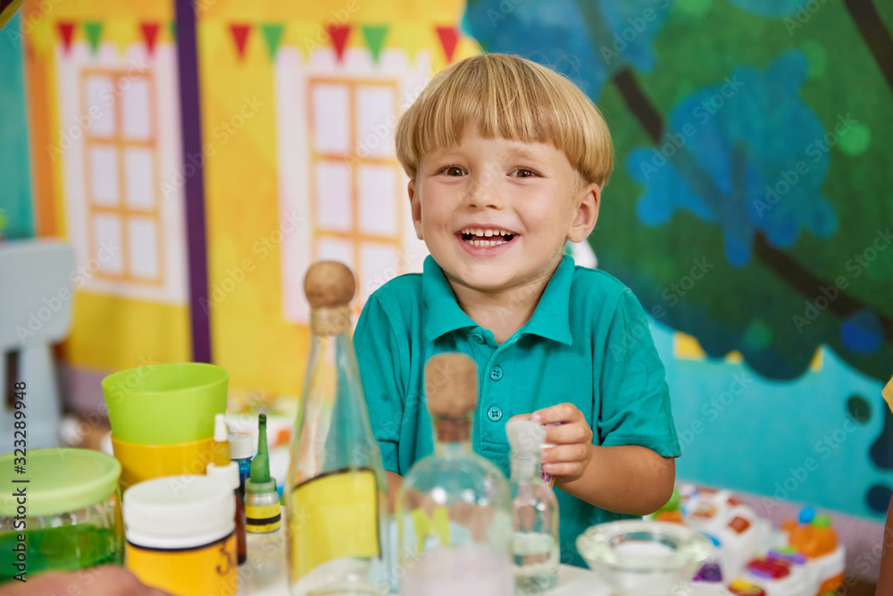Happy smiling kid in playroom of entertainment center. Laboratory glassware flasks on the table. DIY science projects for kids. Happy smiling kid in playroom of entertainment center. Laboratory glassware flasks on the table. DIY science projects for kids.