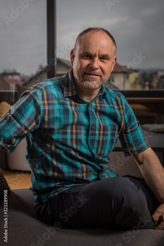 A caucasian man in his late forties with short hair and short gray beard is sitting in a couch and looking into the camera. Forest is seen behind the window. © Anze