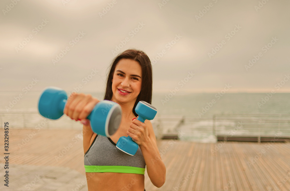 Young cheerful fit woman trains straight arm punch with dumbbells on the beach at sunrise Young cheerful fit woman trains straight arm punch with dumbbells on the beach at sunrise