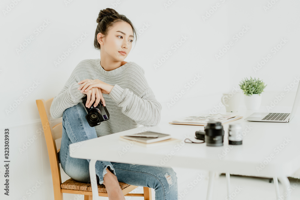 Portrait of young Asian woman photographer with a professional camera sitting on chair and looking away while working at her studio. Freelance Startup Small business owner, Online marketing. SME