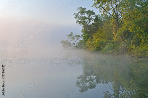 Landscape of the foggy, spring shoreline of Whitford Lake with reflections in calm water, Fort Custer State Park, Michigan, USA