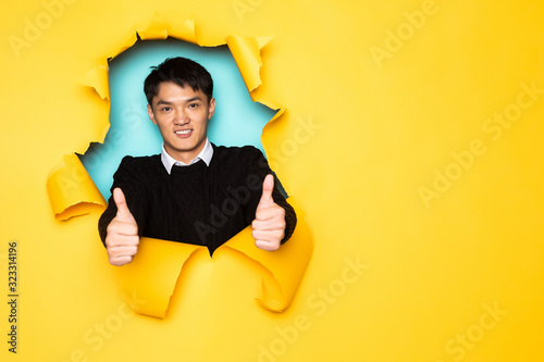 Young chinese man with thumbs up keeps head in hole of torn yellow wall. Male head in ripped paper. © dianagrytsku
