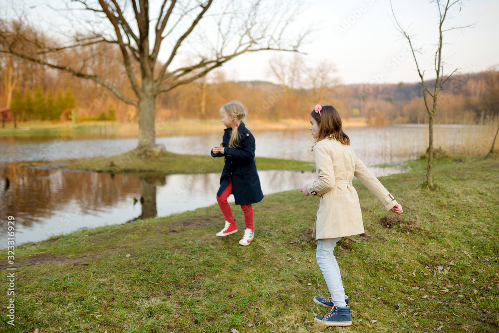 Two cute young sisters having fun by a river on warm spring day. Children playing together by a water. Outdoor family activities in spring. Two cute young sisters having fun by a river on warm spring day. Children playing together by a water. Outdoor family activities in spring.