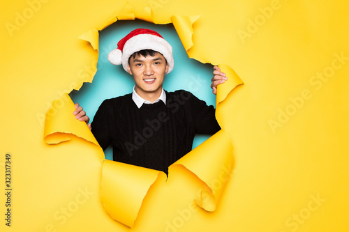 Young chinese man with santa hat keeps head in hole of torn yellow wall. Male head in ripped paper. © dianagrytsku