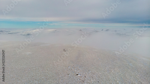 A aerial scenic view of a snowy mountain summit with a mountaineer and mountain range in the background under a majestic blue sky and some altitude white cloud