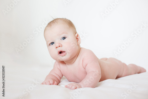 cute naked baby lies on his stomach in bed on a white background. baby with a surprised expression on his face. baby of European appearance. three months to the baby. 