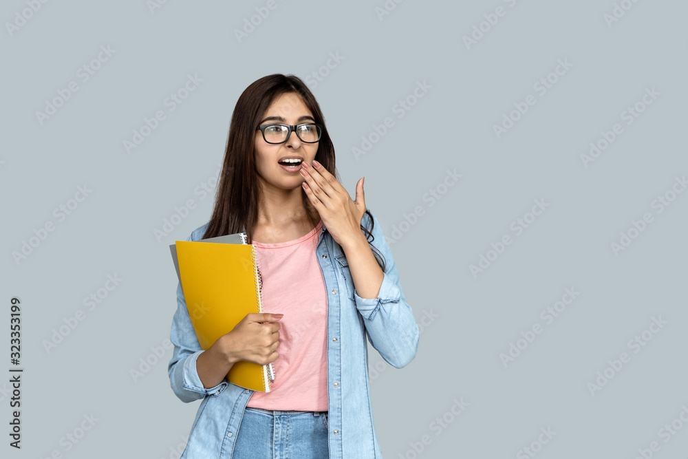 Tired bored young adult indian student girl holding copybooks in hand, looking away yawning and standing isolated on grey background with copy space, boring education concept Tired bored young adult indian student girl holding copybooks in hand, looking away yawning and standing isolated on grey background with copy space, boring education concept