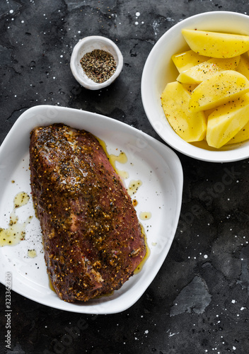 Cooking roasted pork fillet with potatoes and spices - delicious simple lunch on a dark background, top view. Raw ingredients