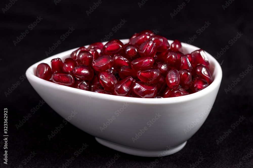 Refined pomegranate: juicy shiny pomegranate seeds are poured into a small bowl and photographed against a black background in the studio. The combination of three colors: red, black and white. Refined pomegranate: juicy shiny pomegranate seeds are poured into a small bowl and photographed against a black background in the studio. The combination of three colors: red, black and white.