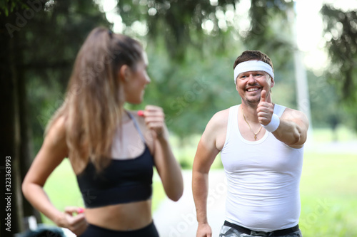 Fototapeta Naklejka Na Ścianę i Meble -  Man in park shows ok gesture to sports girl. Running has beneficial effect on respiratory system body. Happy girl looks at man who smiles at her. Classes relieves stress and uplifting.