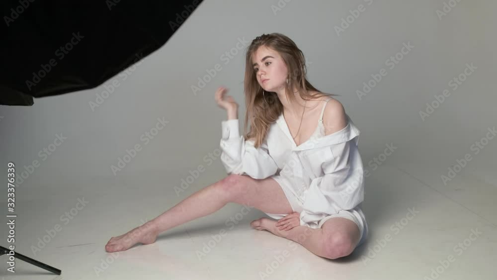 behind the scenes on photo shoot: beautiful young model wearing shirt posing for photographer near softbox on white background, flash lights. model tests of caucasian teen with bare feet in studio