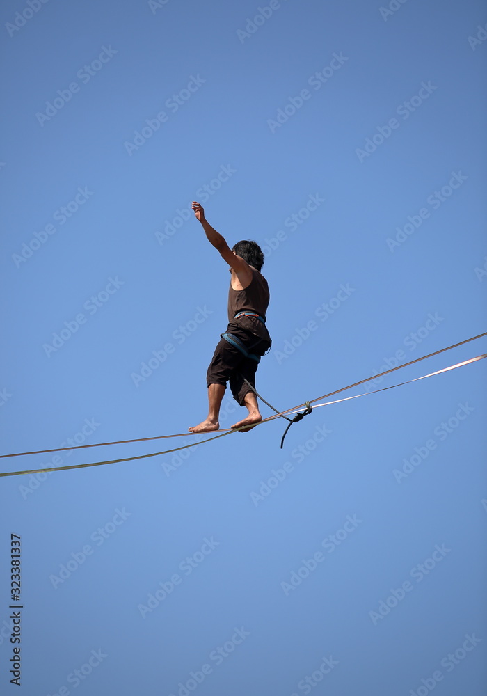 Akrobata spacerujący po linie zawieszonej na dużej wysokości na tle niebieskiego niba - obrazy, fototapety, plakaty 