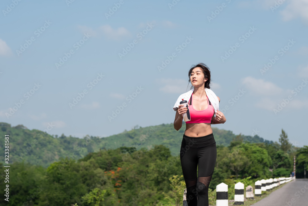 Front view Young asian woman running on road in the morning sunshine outdoors. Female jogger exercising in nature outdoors holding bottled water. Fitness and healthy lifestyle Front view Young asian woman running on road in the morning sunshine outdoors. Female jogger exercising in nature outdoors holding bottled water. Fitness and healthy lifestyle